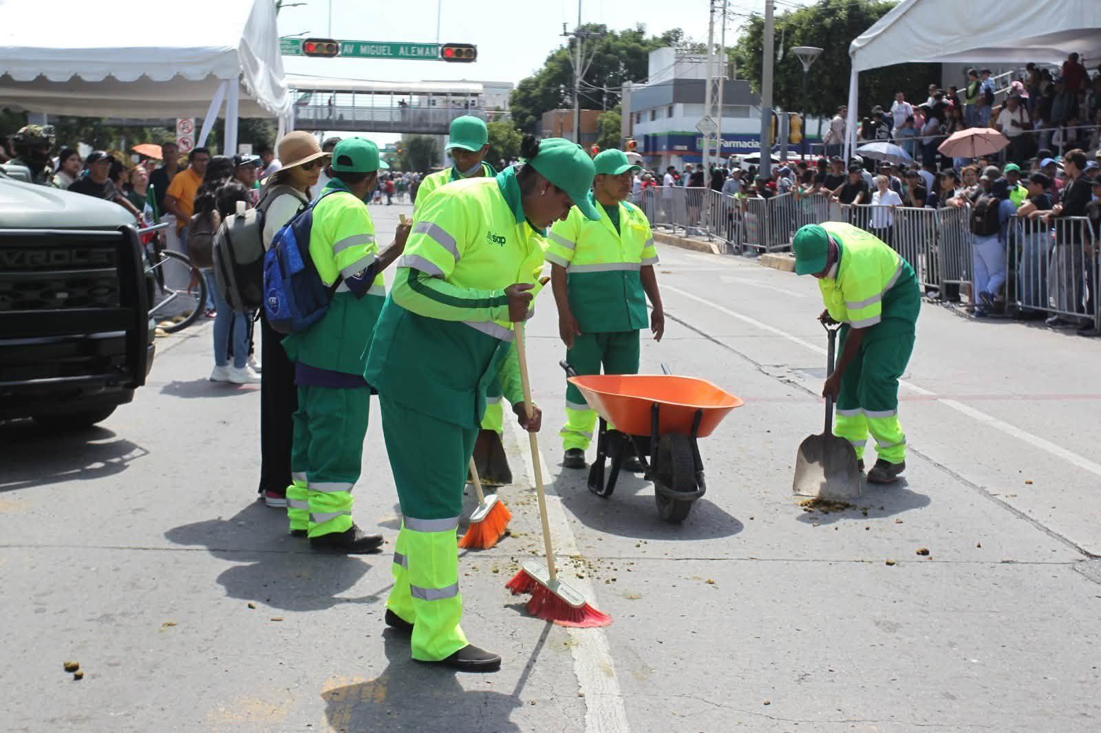 Personal de limpieza con uniforme verde barre una calle del centro mientras la gente mira un evento.