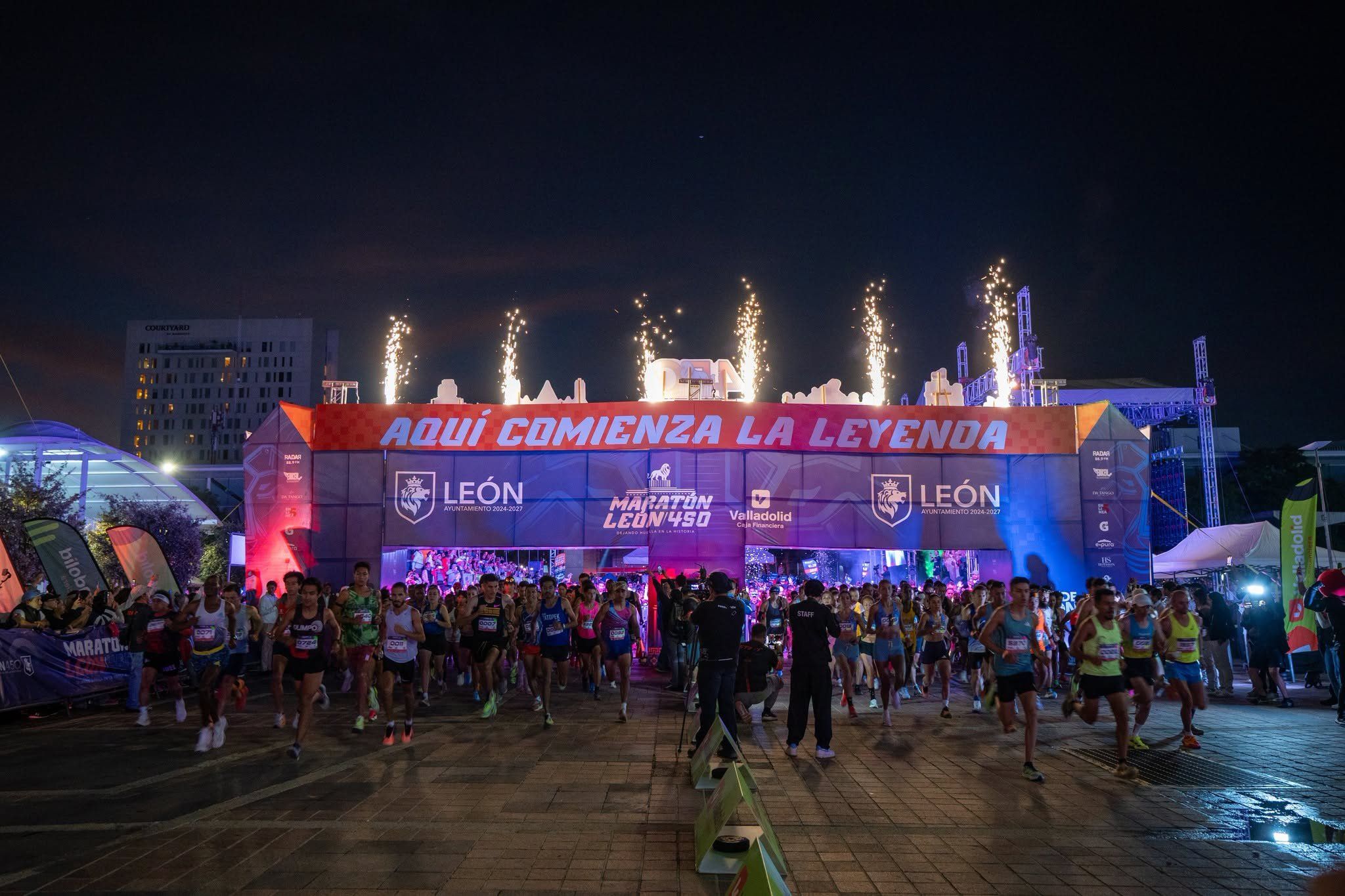 Salida nocturna del Maratón León 450: corredores cruzan el arco con el letrero Aquí comienza la leyenda, entre fuegos artificiales.