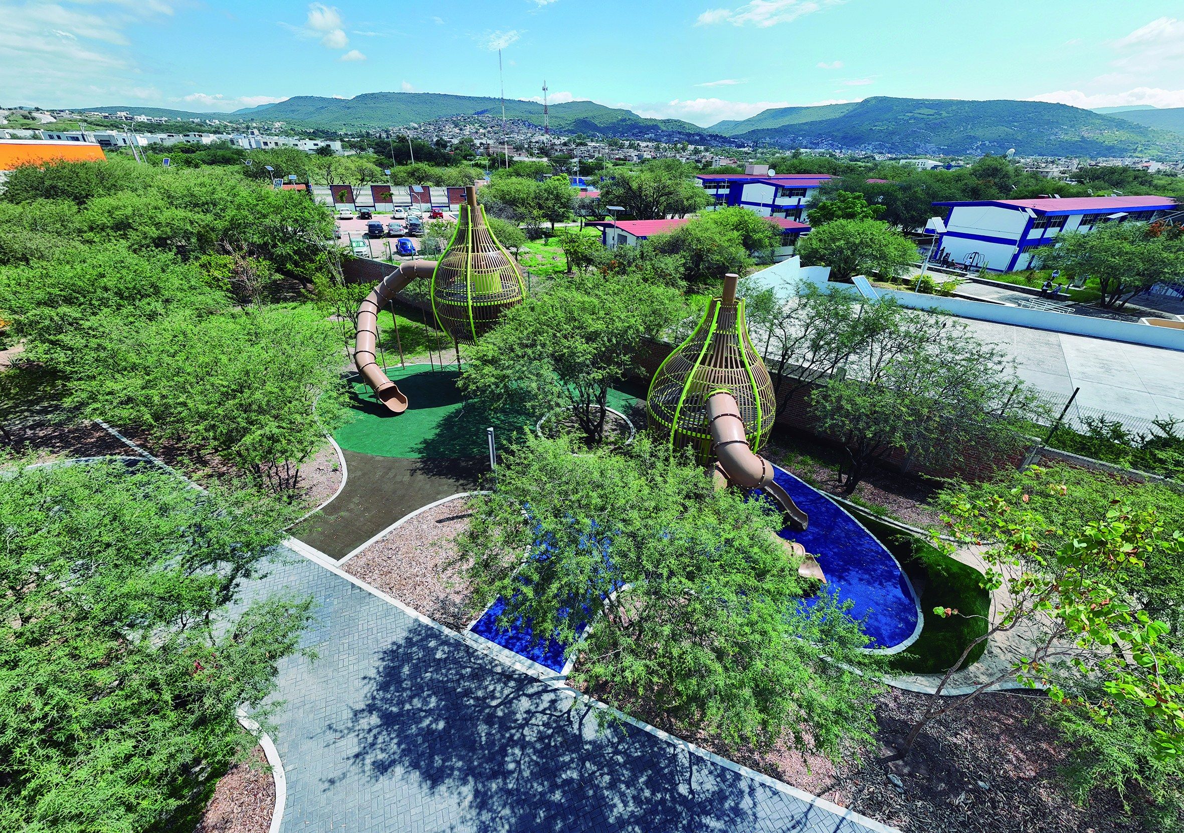 Vista aérea de un parque con juegos infantiles, andadores y árboles, con la ciudad y los cerros al fondo.