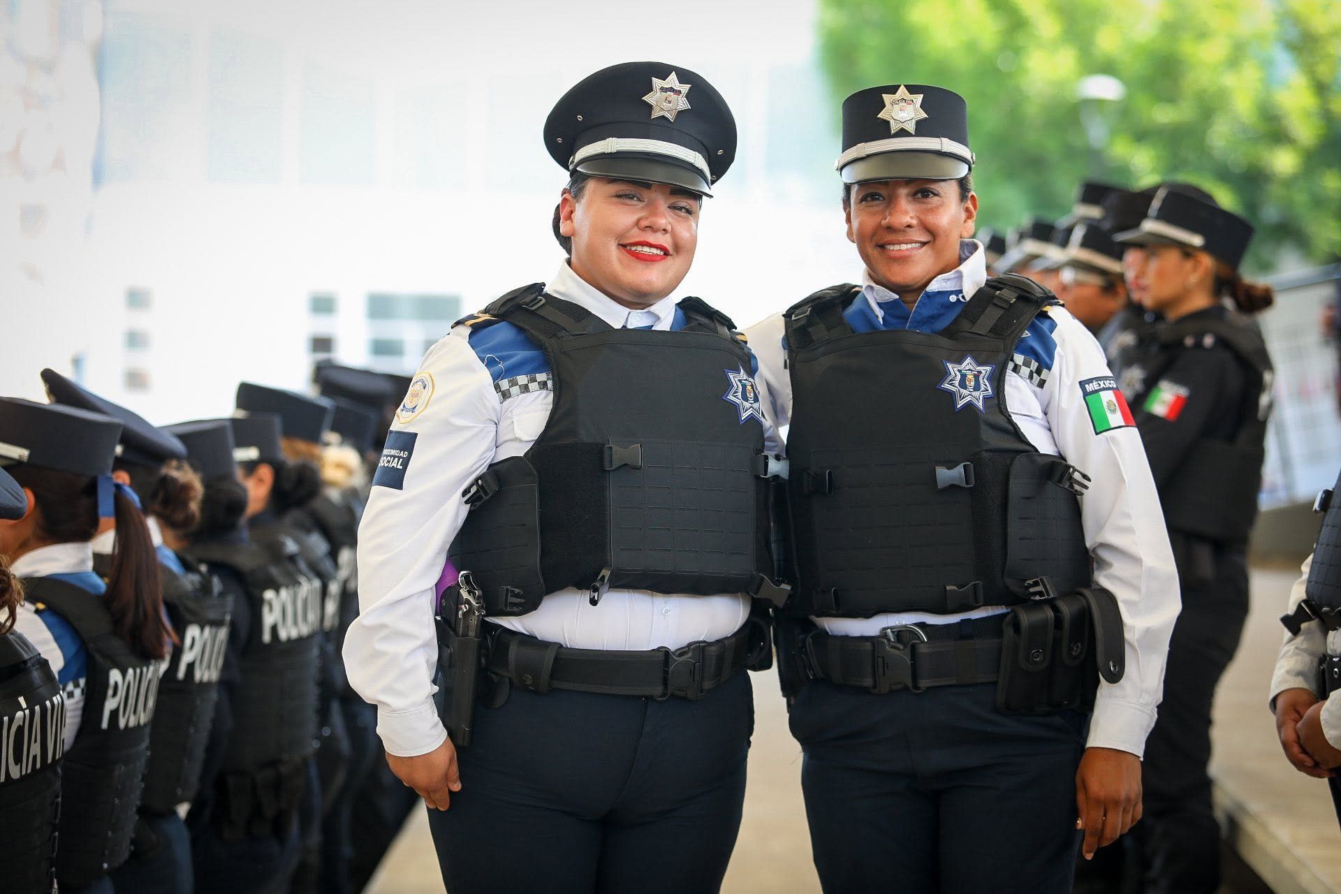 Dos mujeres policías de proximidad sonríen con su uniforme y chaleco, frente a la fila de su generación.
